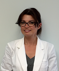 The image shows a woman posing in front of a white wall. She is wearing glasses, a white blazer over a dark top, and has short hair.